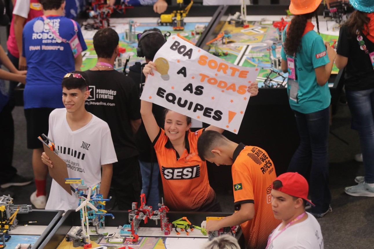 aluno segura cartaz de "boa sorte a todas as equipes" feliz ao redor de outros alunos