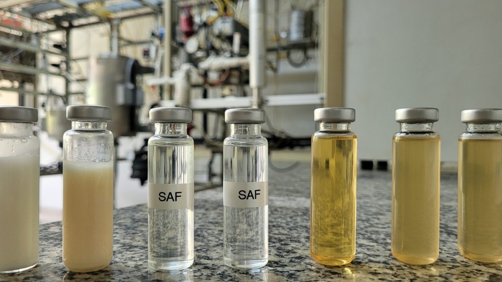 Sete potinhos de vidro enfileirados, com os dois no centro com líquido transparente escrito SAF em bancada de laboratório com fundo desfocado