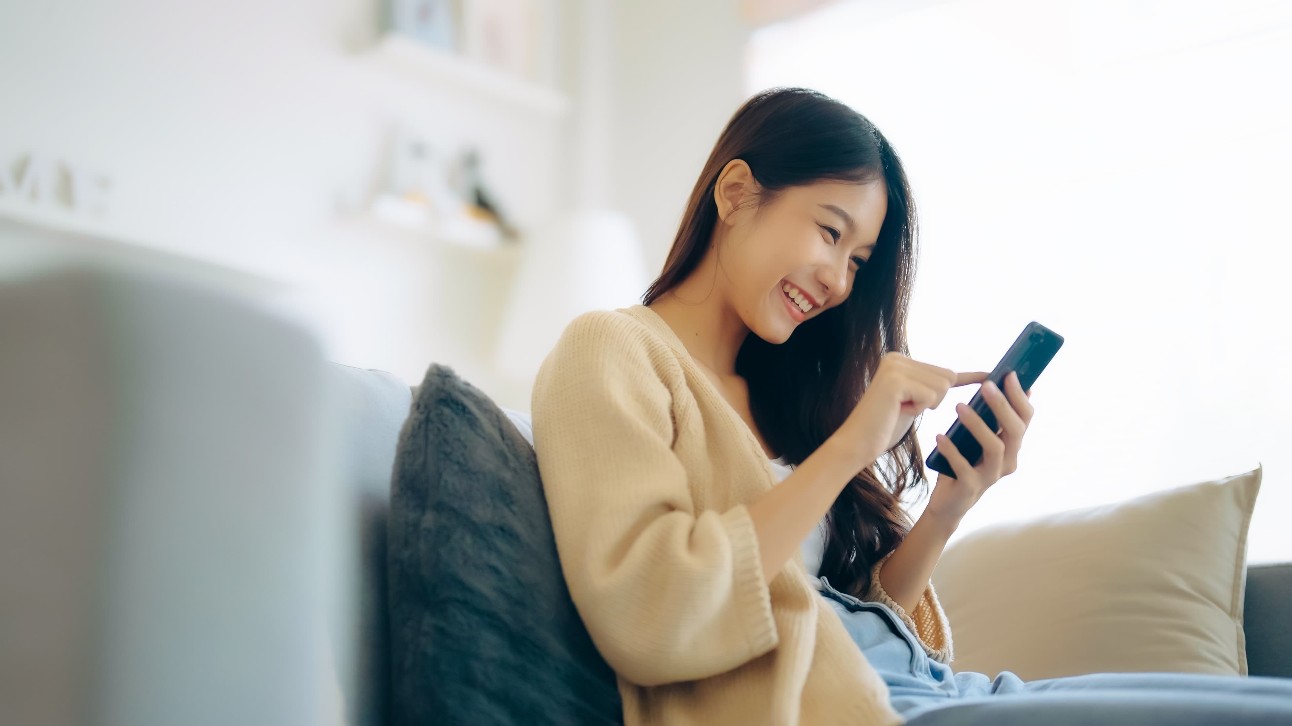 Feliz jovem asiática relaxa no sofá confortável em casa, enviando mensagens de texto no smartphone, garota sorridente usa celular, conversando mensagem on-line, fazendo compras on-line em casa