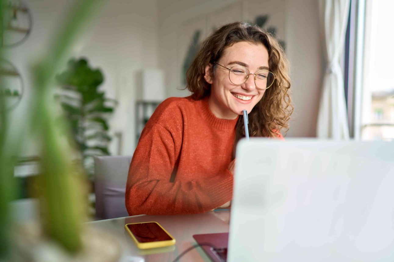 Jovem mulher feliz usando um laptop, sentada à mesa, escrevendo notas enquanto assiste a um webinar, estuda on-line, olha para a tela do computador, aprende aulas on-line ou faz reuniões virtuais por telefone, trabalhando remotamente de casa.