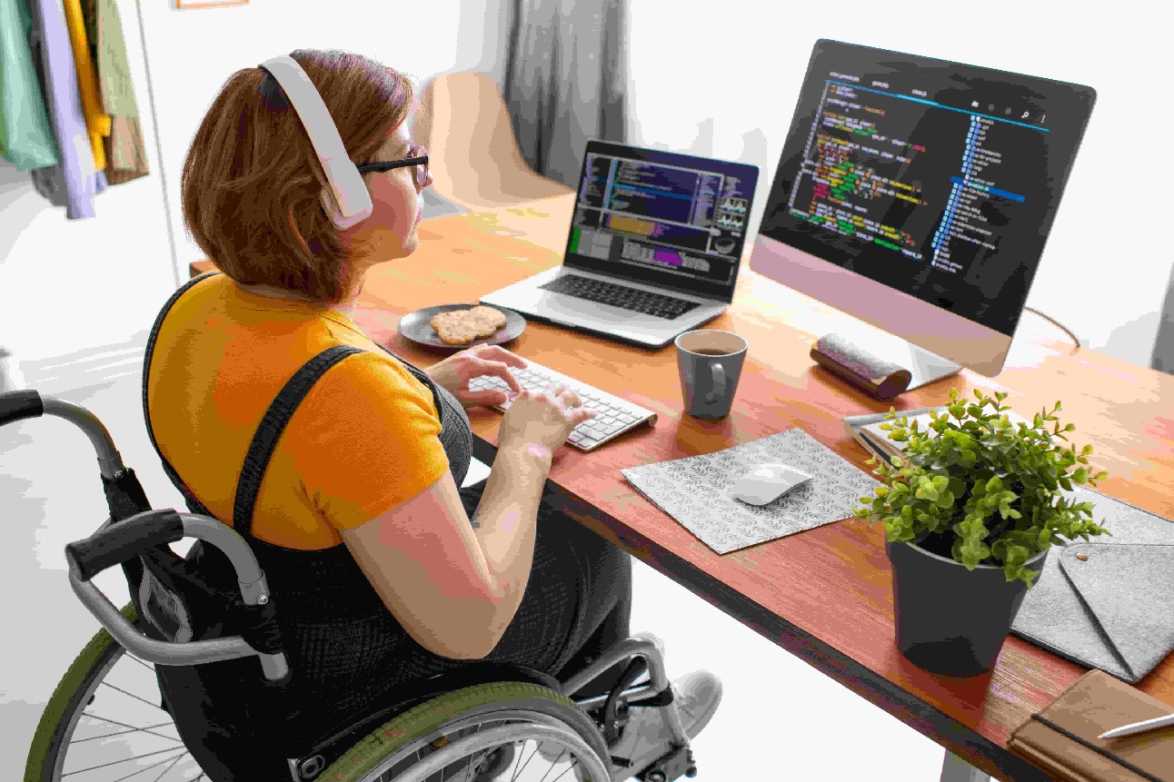 Programadora freelance feminina em fones de ouvido modernos sentada em cadeira de rodas e usando computadores enquanto codifica jogos da web em casa