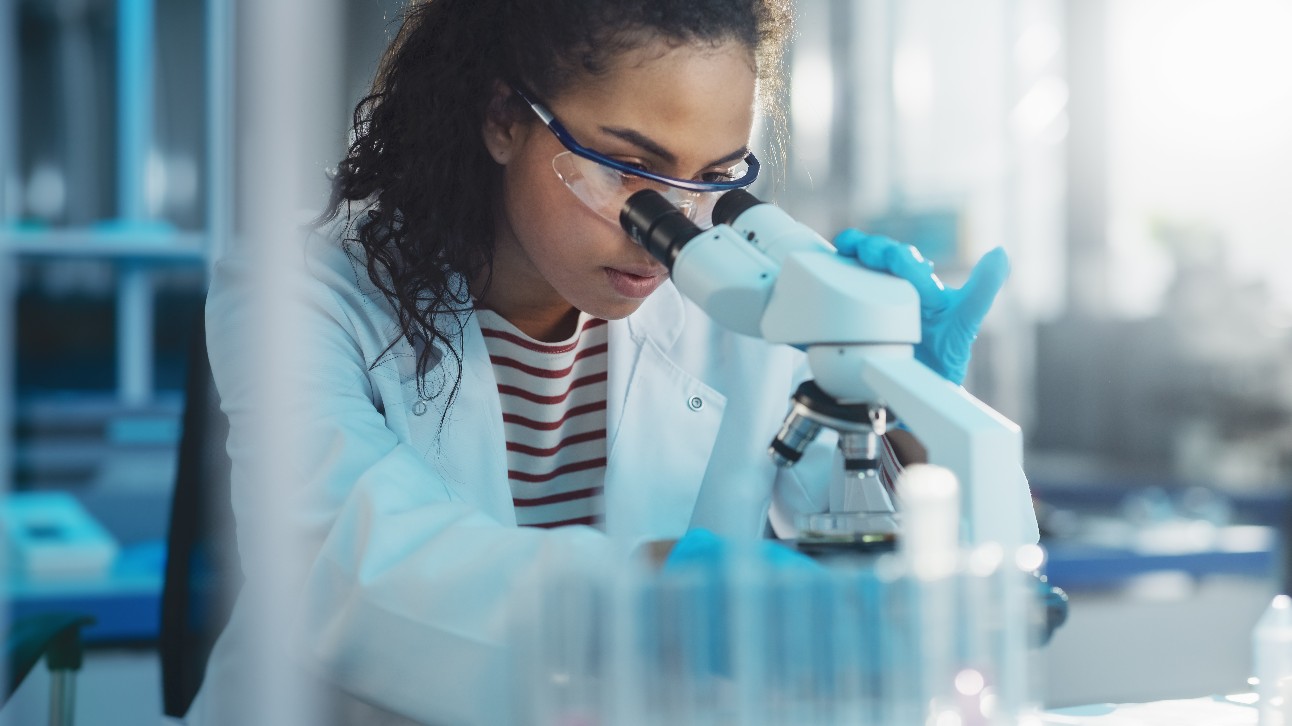 pesquisadora negra, usando óculos de proteção e jaleco branco, analisa amostra em um microscópio