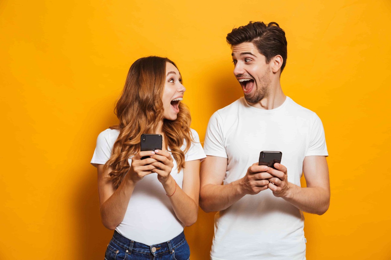 Foto de pessoas excitadas positivas homem e mulher gritando e olhando um para o outro enquanto ambos usando telefones celulares isolados sobre fundo amarelo