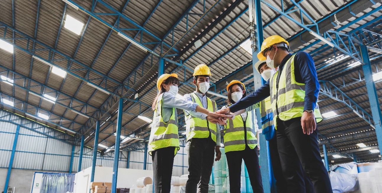 quatro pessoas vestidas com colete, capacete e usando máscara juntam as mãos em sinal de união dentro de uma fábrica