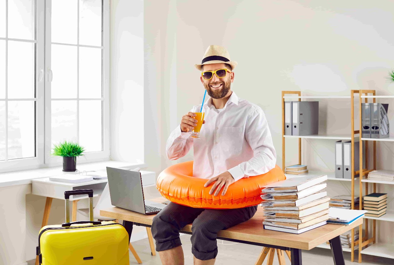 Trabalhador de escritório pronto para ir de férias de verão. Homem sorridente feliz de camisa branca, chapéu de sol, anel de praia e óculos de sol bebe suco de laranja, sentado na mesa com laptop e papéis