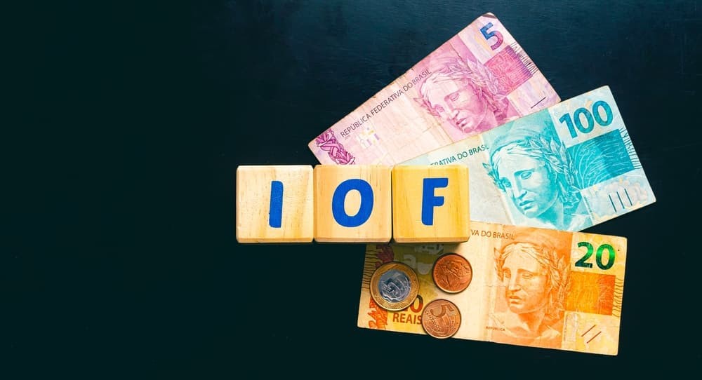 Economia e impostos brasileiros. As iniciais IOF (Imposto sobre Operações Financeiras) escritas em blocos de madeira. Notas de real e algumas moedas na composição da imagem. Vista superior.