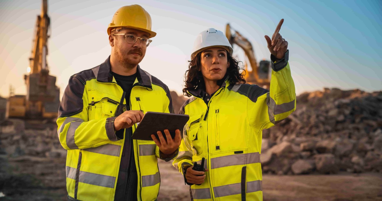 Engenheiro civil caucasiano conversando com uma inspetora hispânica e usando um tablet no canteiro de obras de um novo prédio. Incorporadores imobiliários discutindo negócios, escavadeiras trabalhando.