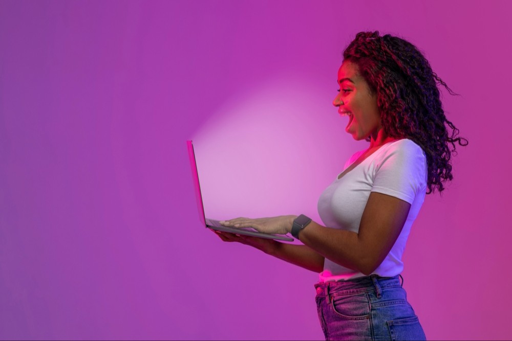 Mulher surpresa olhando para a tela brilhante do laptop, jovem reagindo emocionalmente à oferta online enquanto está sob luz neon sobre fundo roxo, vista lateral