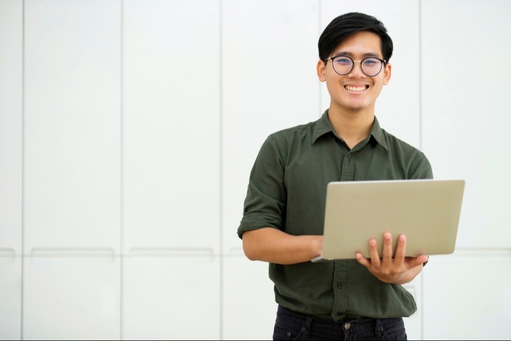 homem jovem asiático, usando óculos de grau, veste camisa social verde escura e segura um notebook.