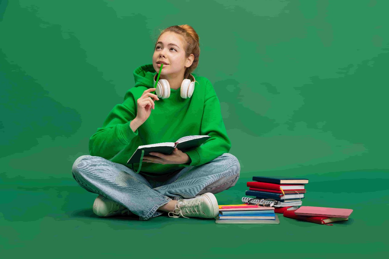 Retrato de jovem, estudante em pano casual, sentada no chão com expressão pensativa, estudando isolada sobre fundo verde do estúdio.