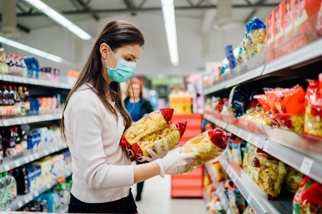 mulher branca, com cabelo liso, máscara, blusa de manga comprida, lê embalagem de pacote de macarrão em supermercado