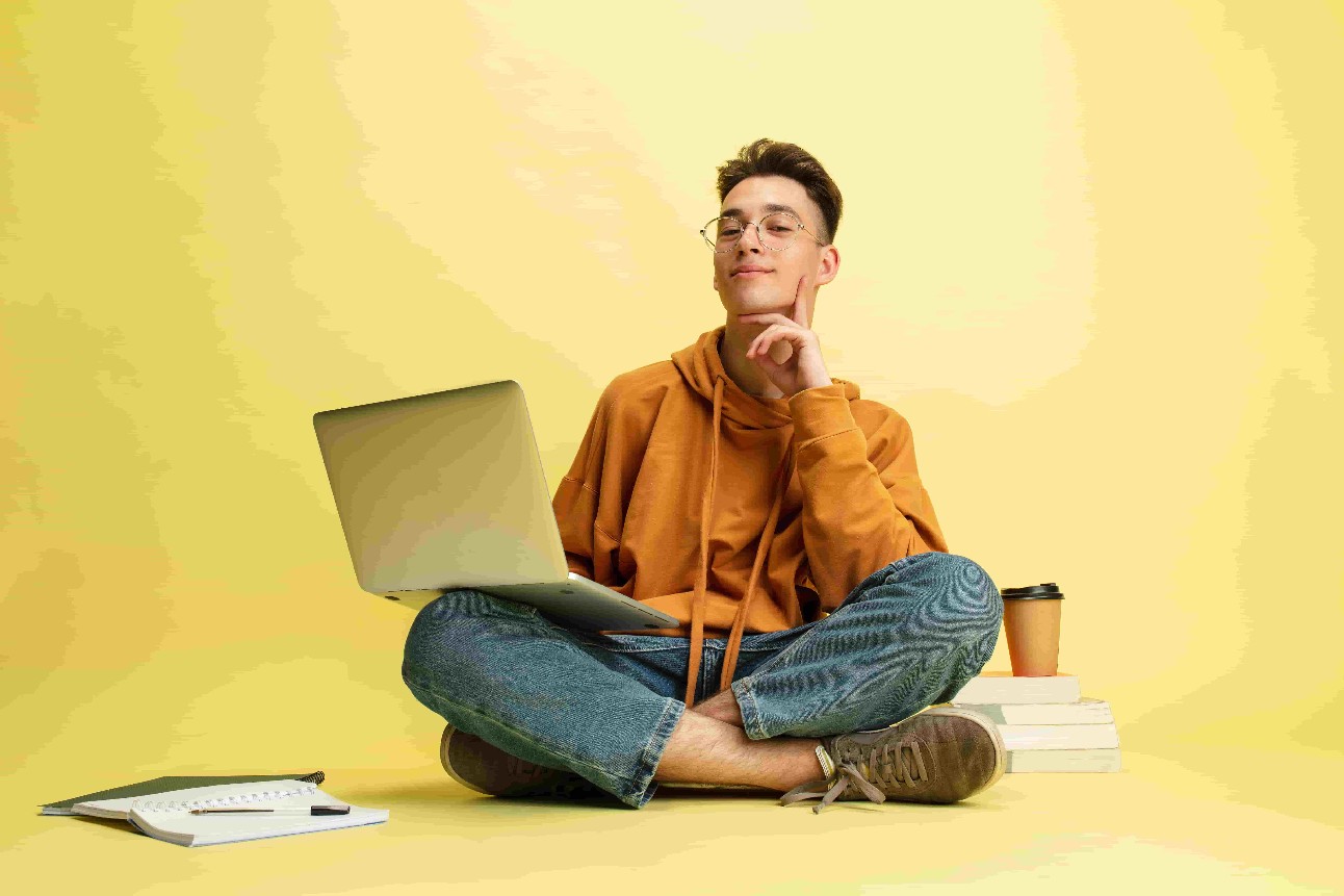 Estudando, fazendo lição de casa. Um jovem homem branco sorridente, estudante de óculos, senta-se no chão com o laptop no fundo amarelo do estúdio.