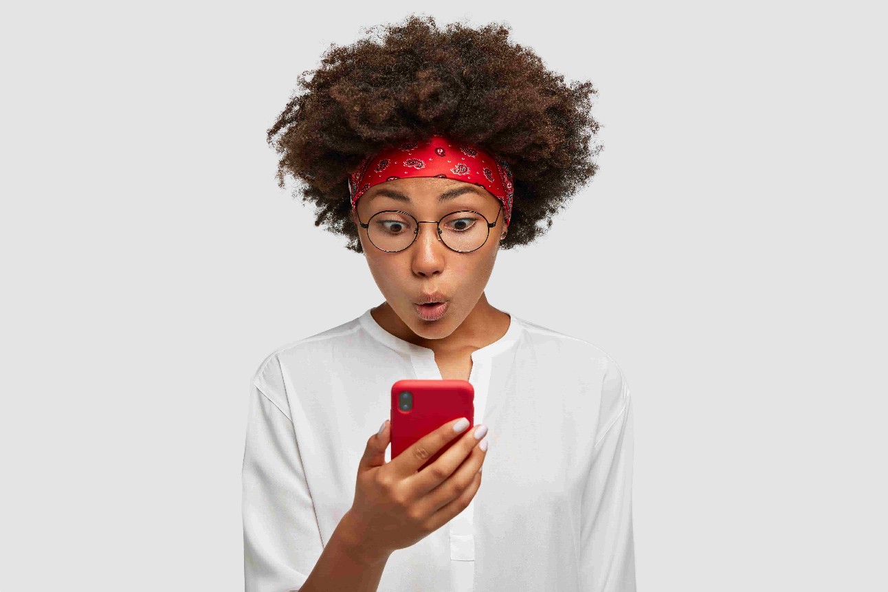Mulher preta de black power, usando blusa branca, óculos e faixa da cabelo vermelha, olha impressionada para celular.