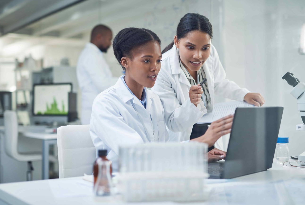 Duas mulheres, uma branca e outra negra, em laboratório com computador e jalecos brancos. Homem negro ao fundo com jaleco branco
