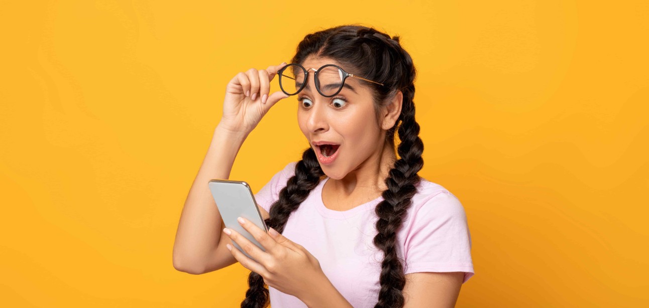 Uau, ótimas notícias. Retrato de uma mulher indiana surpresa e animada segurando e usando um smartphone, navegando nas redes sociais. Jovem emocionada tendo sucesso, tirando os óculos, fundo de estúdio amarelo.