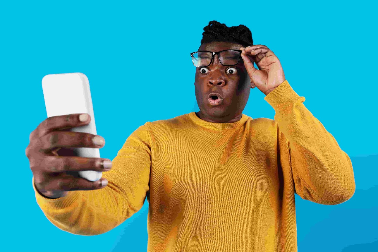 Oferta incrível. Retrato de um homem negro chocado olhando para a tela do smartphone, um jovem afro-americano surpreso tirando os óculos, lendo uma mensagem inesperada em pé sobre um fundo azul