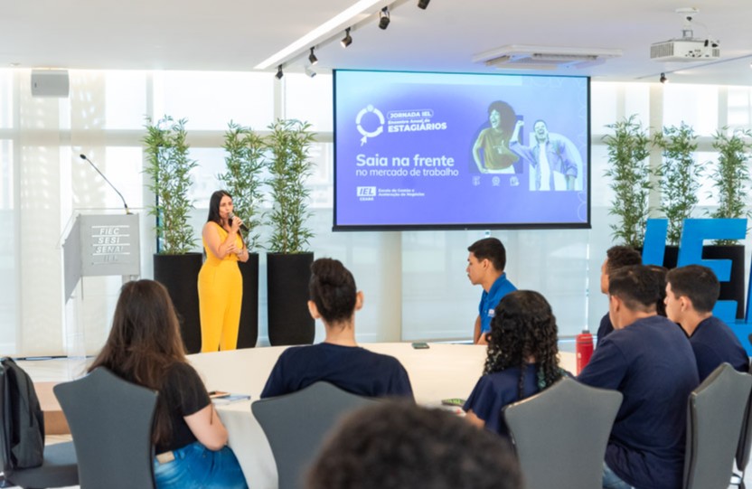 mulher palestra em meio a grupo de pessoas, a mulher é branca, usa macacão amarelo e tem cabelo castanho