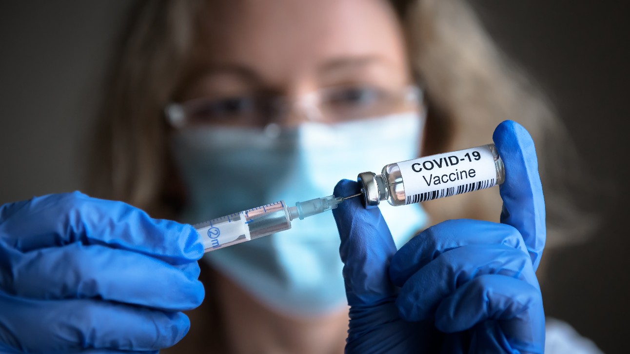Mulher branca, de máscara azul, manipula seringa e uma dose da vacina contra covid-19