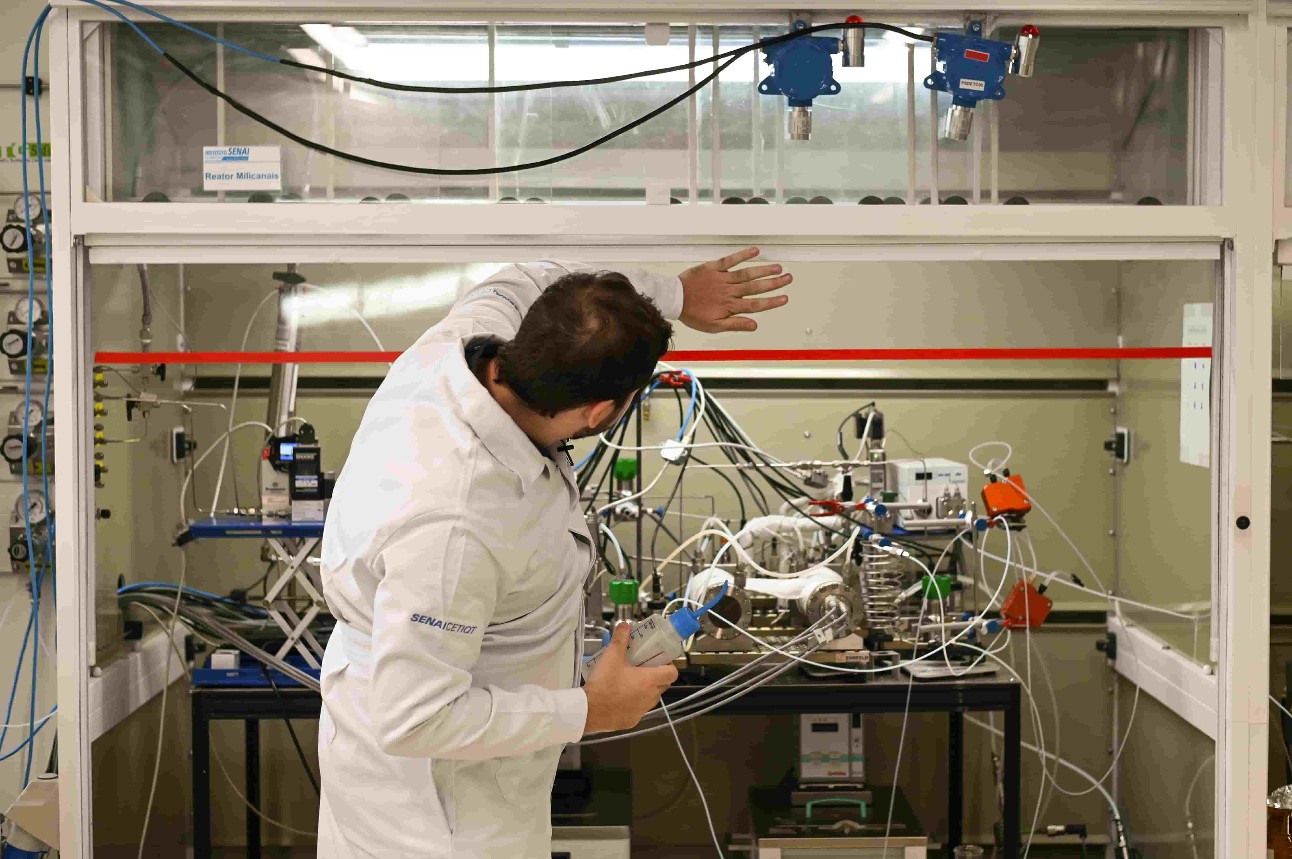 Homem, de jaleco, está de costas mostrando máquina com vários fios em laboratório.