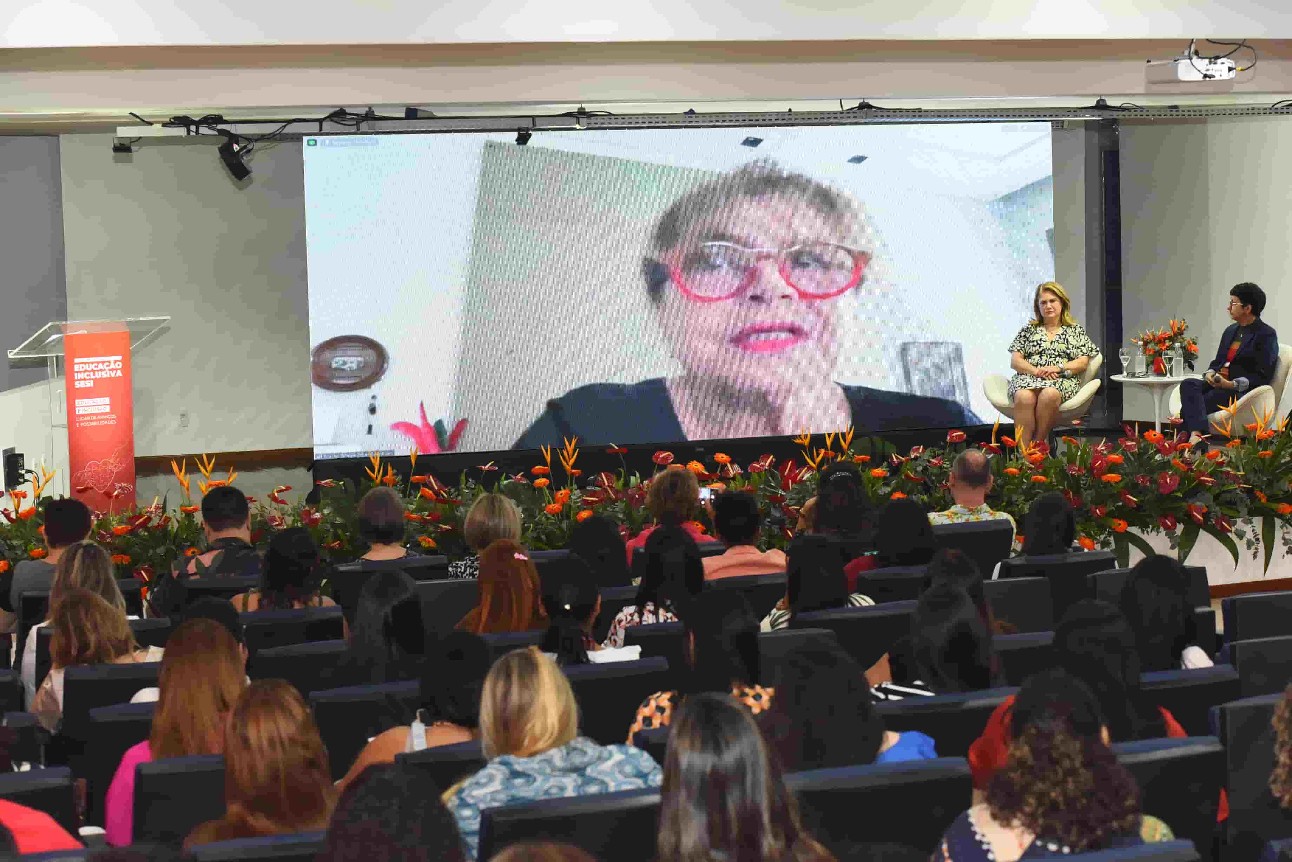 Foto de palestra com várias pessoas sentadas assistindo um telão, com o rosto de uma mulher branca de óculos vermelho