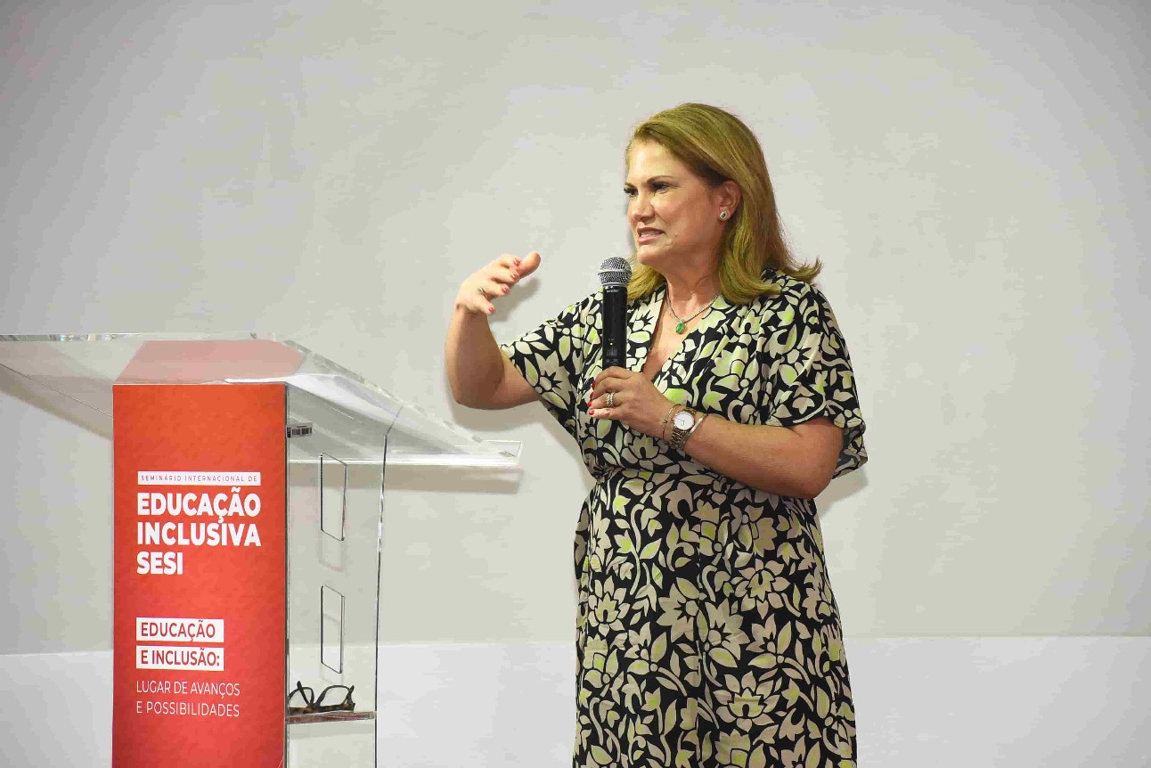 mulher branca, loira, de vestido florido, fala ao microfone e em frente está um púlpito