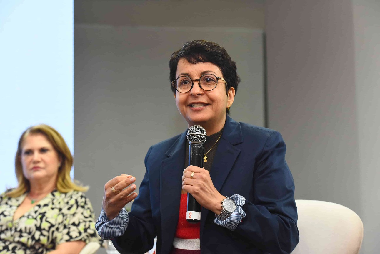 mulher branca de cabelos curtos e castanhos, usa óculos, está sentada falando ao microfone