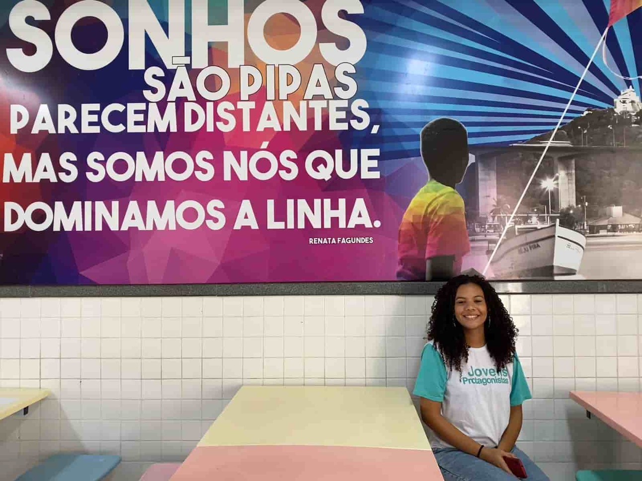 Aluna adolescente preta, de cabelos cacheados e sorriso largo, está sentada ao lado de uma mesa. Atrás tem um painel colorido.