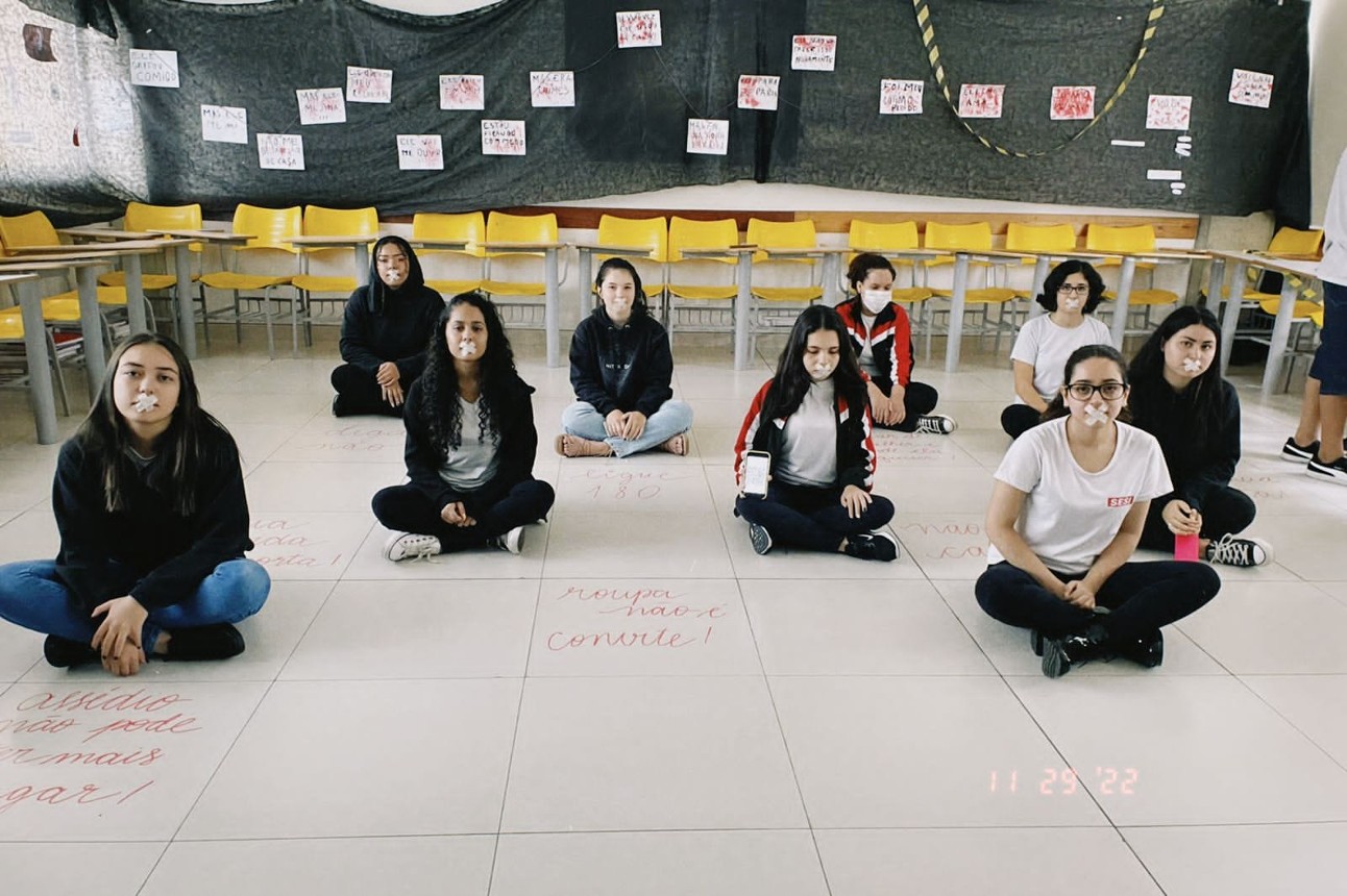 Nove estudantes sentadas com papéis em forma de X na boca. No chão e ao fundo da sala de aula, há mensagens e cartazes
