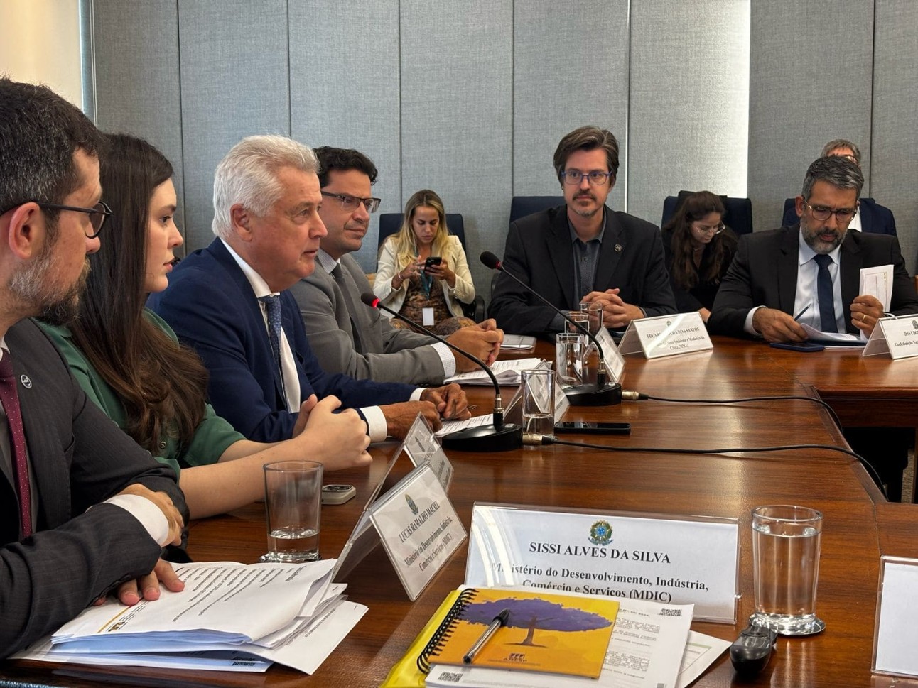 imagem colorida de pessoas reunidas em volta de uma mesa, com vários papéis e algumas placas que descrevem os nomes das pessoas