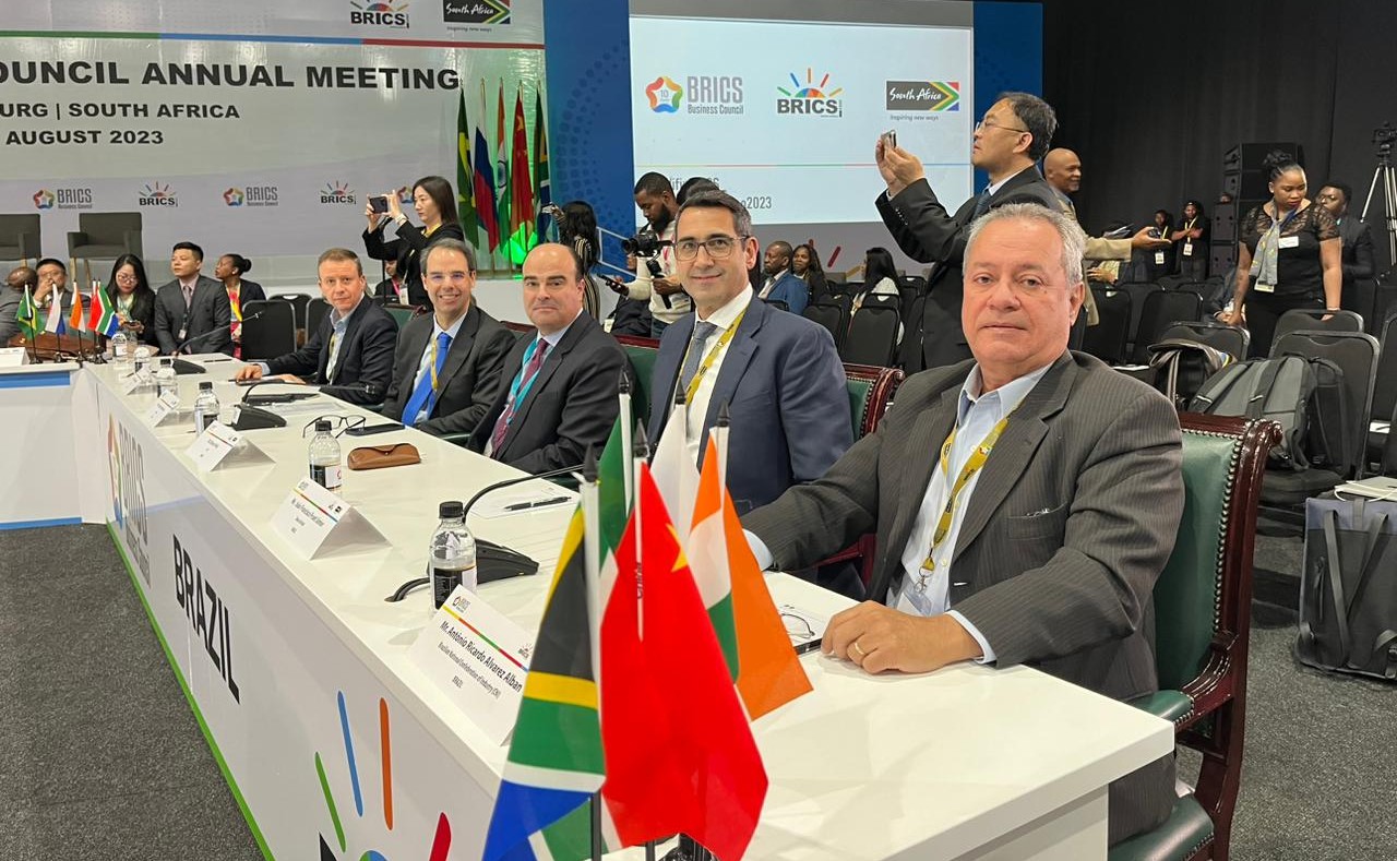 Homens de terno sentados em mesa com toalha branca e bandeiras dos países do Brics em cenário de conferência