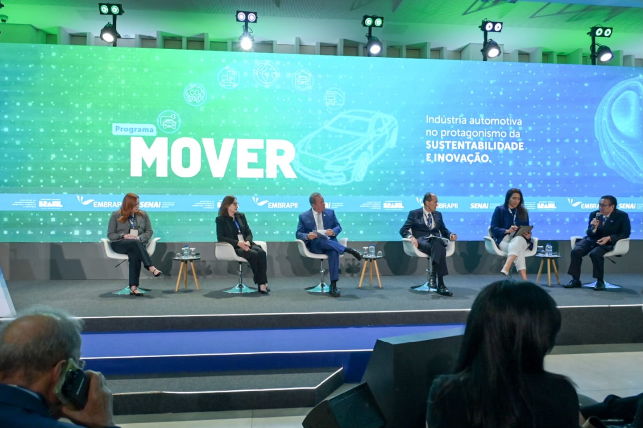 No palco, seis pessoas — quatro homens e duas mulheres — estão sentadas em poltronas brancas, participando de uma conversa mediada, com copos d’água e materiais de apoio sobre mesas pequenas ao lado.