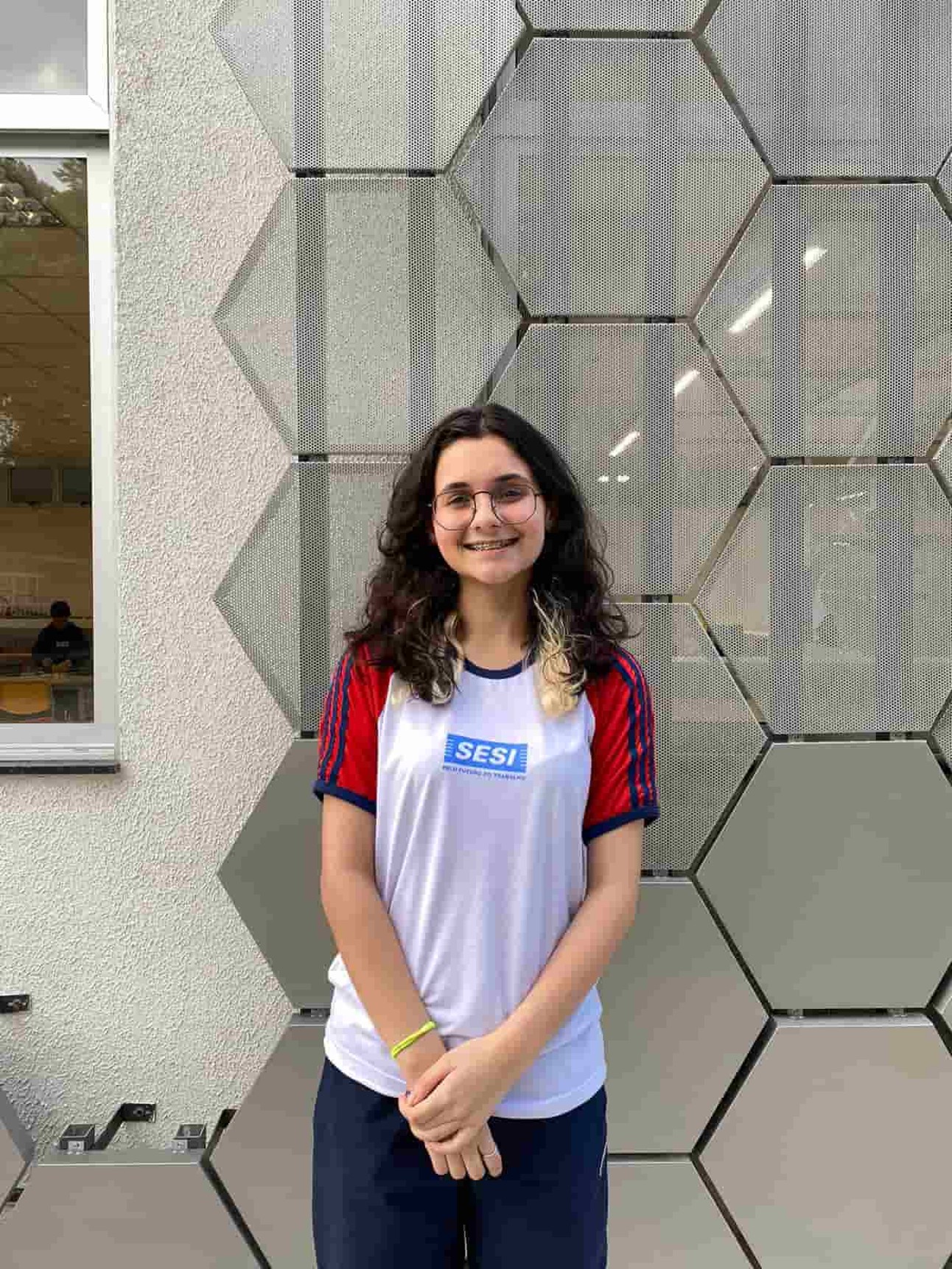 estudante, de cabelos lisos e uniforme do SESI, posa em frente a parede com sorriso tímido e mão cruzadas em frente ao corpo.