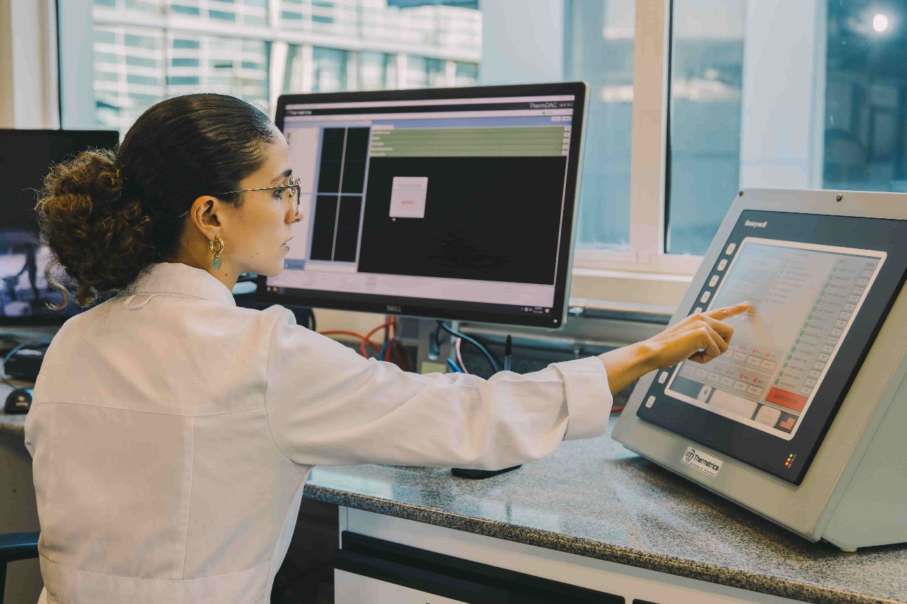 Mulher branca sentada em laboratório em frente a uma tela de computador apontando para equipamento ao lado